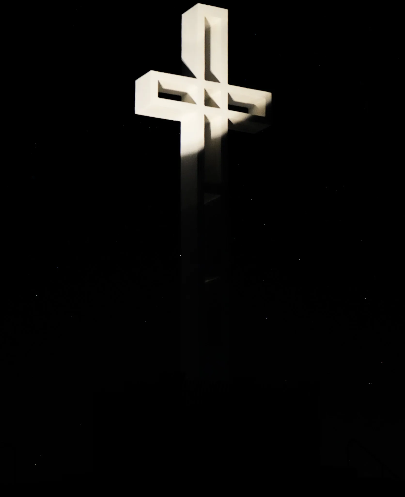 A tall white cross monument floodlit from below against a black night sky, its two crossbars throwing hard shadows down the shaft. Nothing else in the frame is lit.