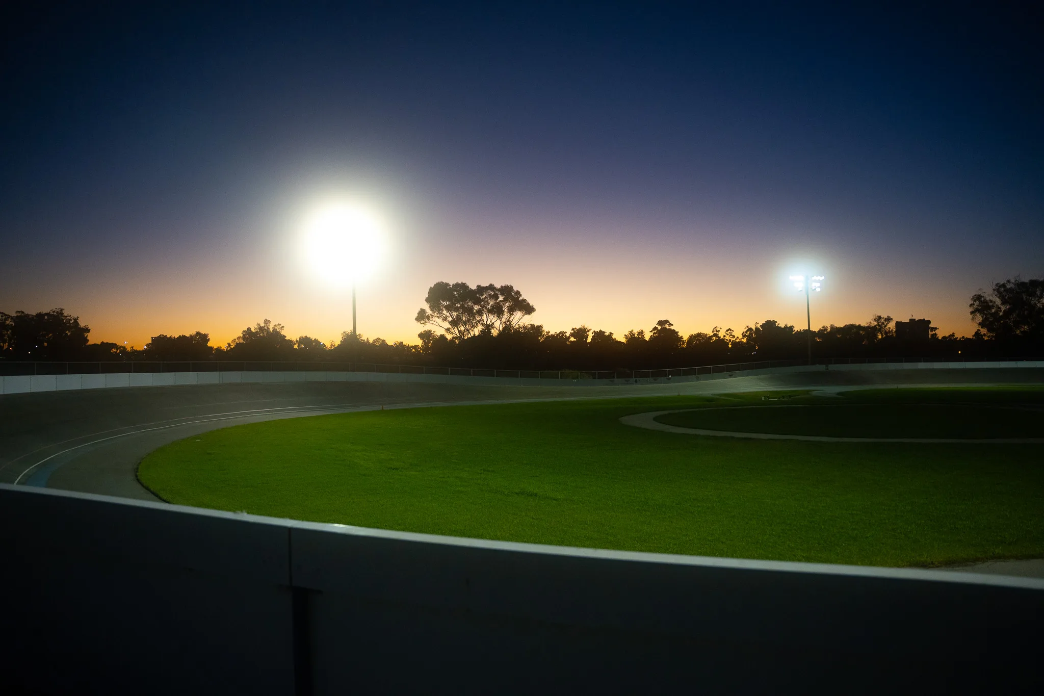 The same velodrome at dusk, empty: floodlights burning over a treeline against an orange and deep-blue sky, the banked track sweeping around a lit green infield.