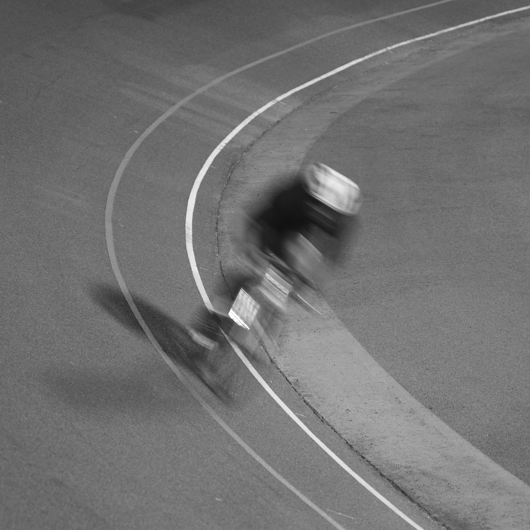 A black-and-white frame of a cyclist carrying speed through the banking of a velodrome, blurred to a smear of dark shapes by a long exposure. The painted sprinters’ lines curve cleanly through the grey concrete around him.
