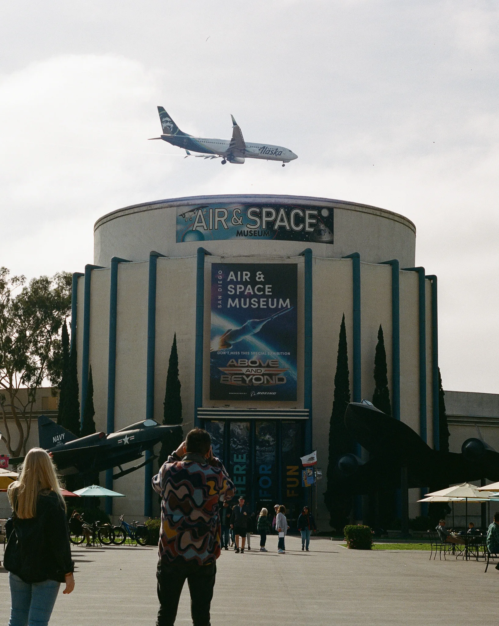 An airliner passing low over the drum-shaped facade of the San Diego Air and Space Museum, whose signage and exhibition banner are legible. A jet fighter and a dark aircraft are mounted either side of the entrance; two people watch from the foreground.