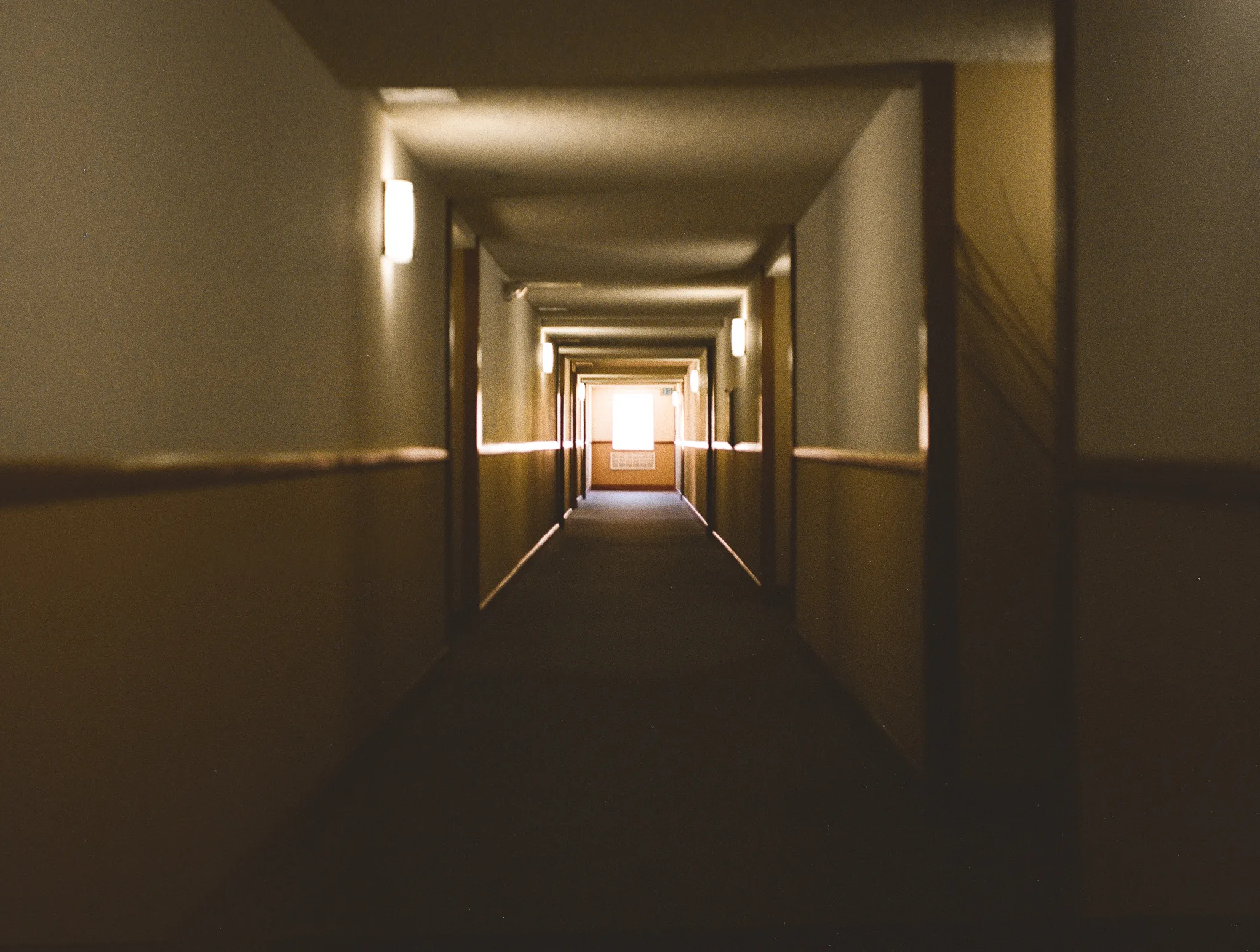 A dim residential corridor in warm tungsten light, wall sconces repeating down both sides and a bright window at the far end. The near half of the frame falls away into shadow.