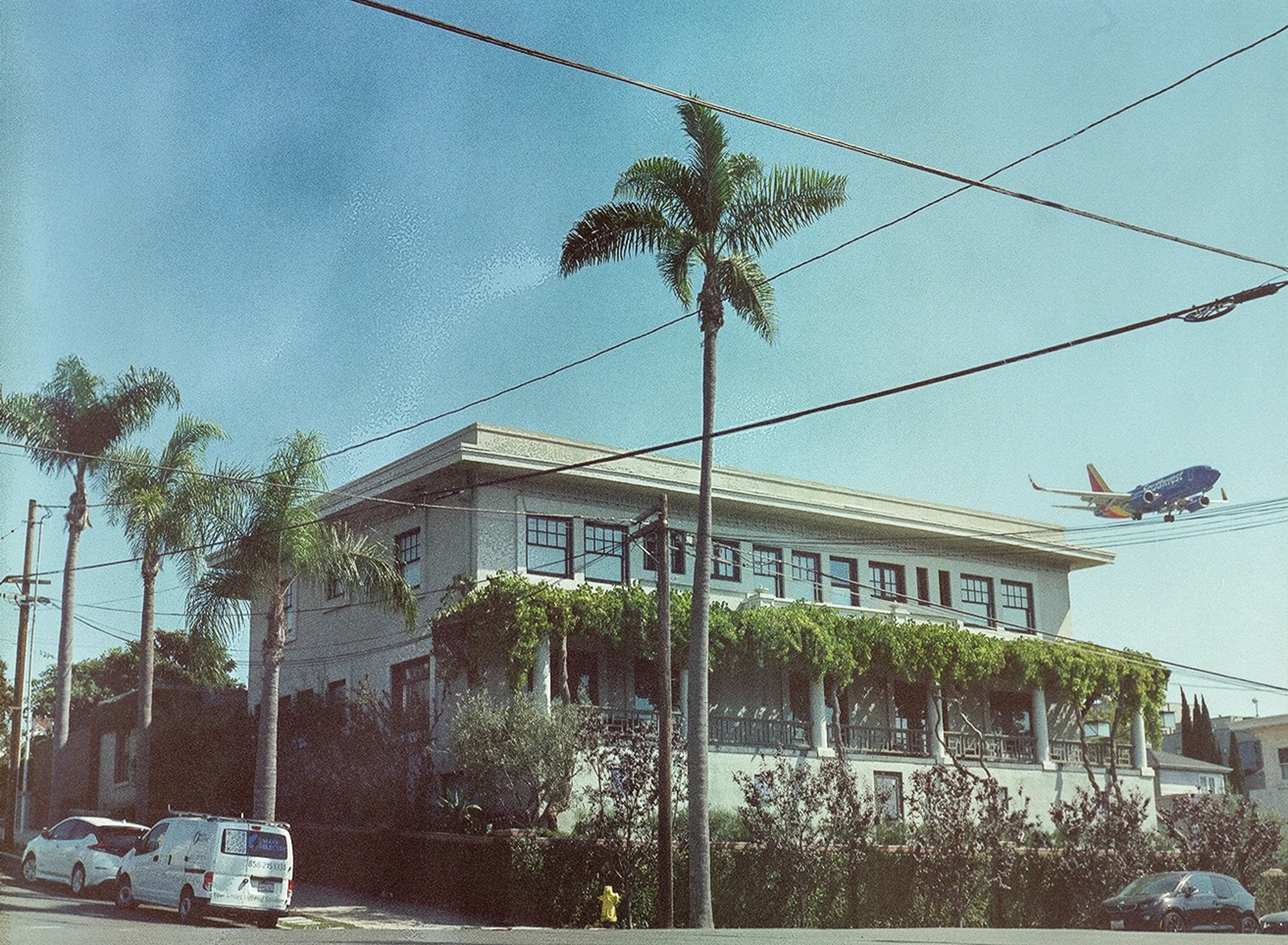 A passenger jet on final approach, low over a two-storey stucco building whose balconies are curtained in climbing vine. Palms and overhead power lines cross a hazy blue sky.