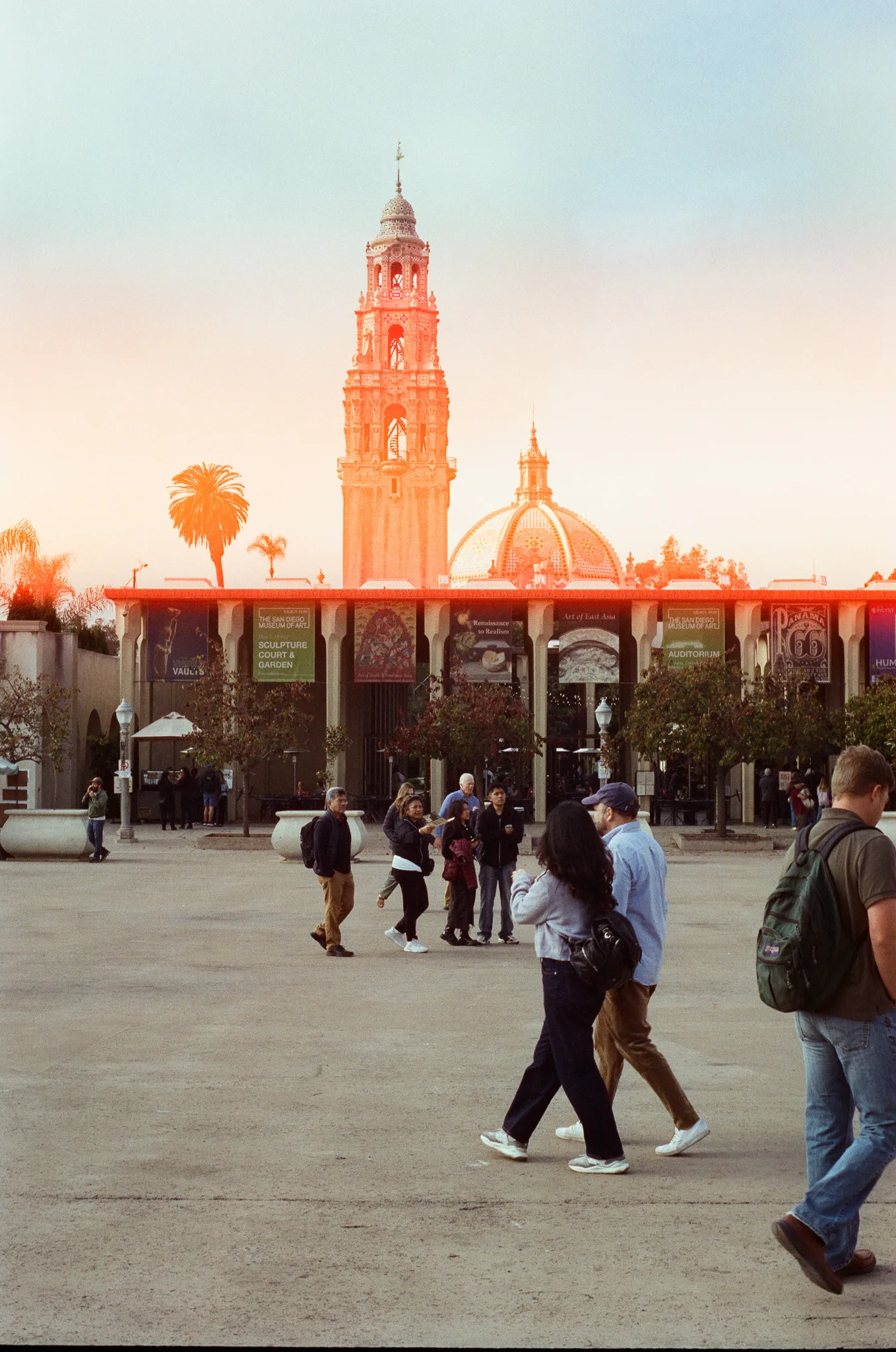 An ornate Spanish Colonial bell tower and tiled dome lit orange by low sun, above a long arcade hung with banners. People cross the open plaza in front of it in silhouette.
