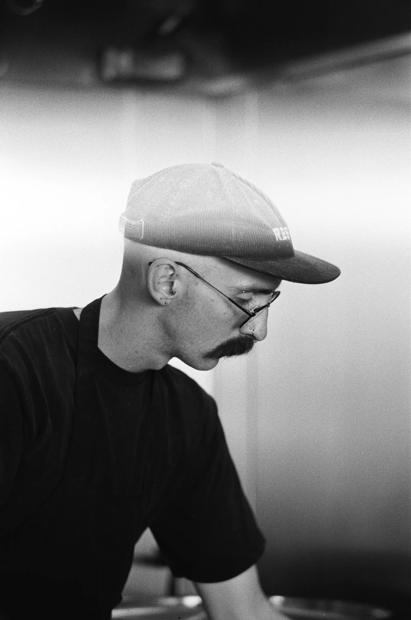 A black-and-white portrait of a man in a light cap, wire-rimmed glasses and a black t-shirt, head lowered and turned in profile as he works at something out of frame. Brushed steel panelling behind him.