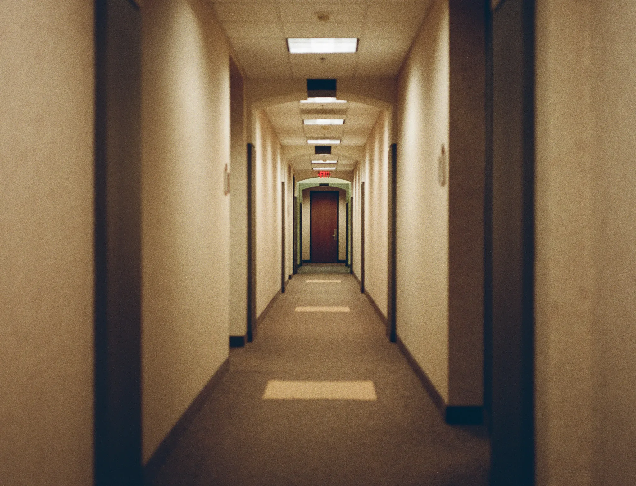 A long, empty office corridor under recessed fluorescent panels, the pale walls and dark doorframes converging on a closed wooden door and a small red exit sign at the far end.