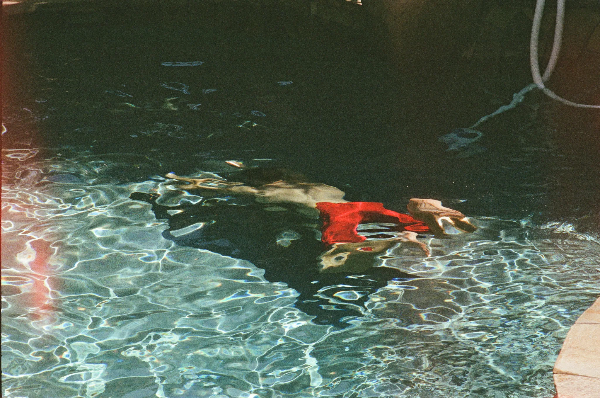 Someone submerged just under the surface of a swimming pool, seen from the edge. A red garment and a pale hand read clearly through the water; the rest of the figure breaks apart in the refraction and the surface glare.